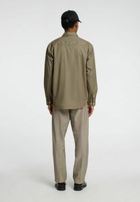 Camicia khaki a maniche lunghe con una vestibilità rilassata, dettagli con bottoni e una consistenza liscia, abbinata a pantaloni grigi chiari e scarpe nere.