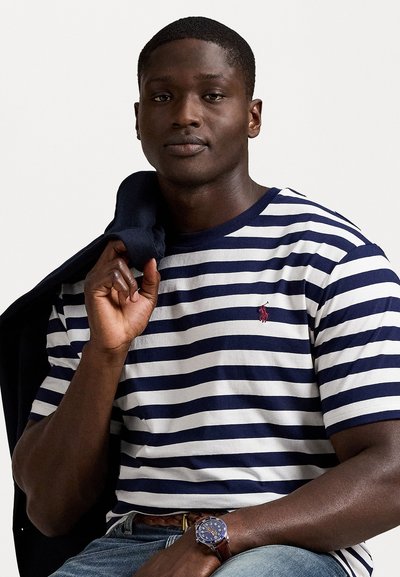 Jūras spēku un baltu svītru T-krekls ar apaļu kakla izgriezumu, uz kura ir neliels sarkans logos. Uzvilkts ar žaketi, kas izmesta uz viena pleca.