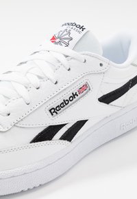 Biele kožené tenisky s čiernymi akcentmi, s nápisom Reebok a textúrovanou gumovou podrážkou. Obsahujú biele šnúrky a polstrovaný jazyk.