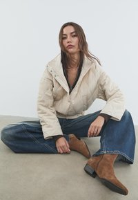 Giacca trapuntata beige con colletto alto e chiusure a pressione, abbinata a jeans blu larghi e stivaletti in camoscio marrone con tacco.