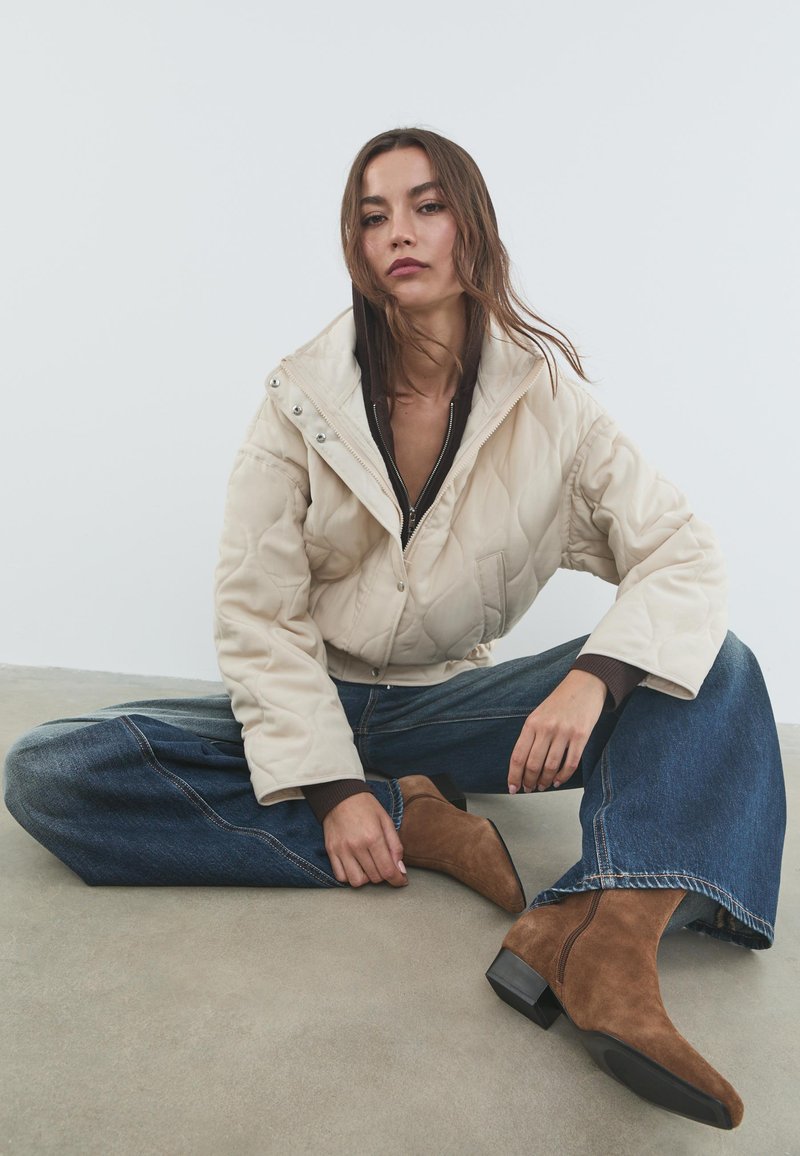 Giacca trapuntata beige con colletto alto e chiusure a pressione, abbinata a jeans blu larghi e stivaletti in camoscio marrone con tacco.