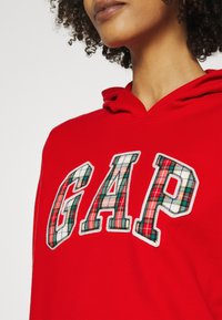 Sudadera con capucha roja con un gran logo "GAP" en patrón de cuadros con costuras blancas. Tela suave y capucha ajustable con cordón.