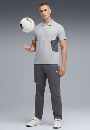 Hellgraues Polohemd mit einer dunkelgrauen Tasche, akzentuiert mit neongrün; kombiniert mit dunkelgrauen Hosen mit einem weißen Seitenstreifen. Hält einen Fußball.
