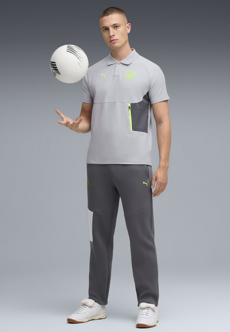 Svetlo šedé polo s tmavosivou náprsní kapsou, akcentované neonovo zelenou; kombinované s tmavosivými nohavicami s bielym bočným pruhom. Drží futbalovú loptu.