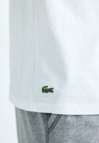 T-shirt en coton blanc avec un petit logo de crocodile vert sur l'ourlet, associé à un pantalon gris à taille élastique.