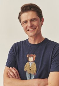 Tumši zila T-krekls ar attēlu, kurā redzams lācis bēšā džemperī un zilās džinsās, valkāts uz modeļa ar sakrustotām rokām. Gluda auduma tekstūra.