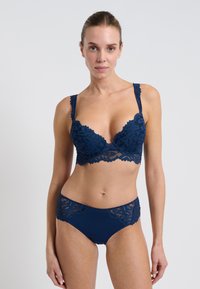 Námořnická modrá souprava spodního prádla obsahující krajkový bralet s vlnitými okraji a odpovídající kalhotky s krajkovými detaily, které zvýrazňují složité květinové vzory.