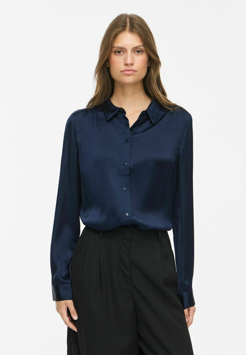 Femme portant un chemisier en satin bleu marine à manches longues, rentré dans un pantalon noir taille haute, debout devant un fond blanc uni.
