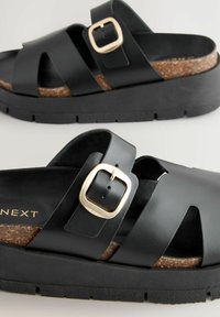 Sandali platform neri con fondo in sughero, caratterizzati da ampie cinghie incrociate e un accentuato bottone dorato. Suola in gomma testurizzata per una maggiore aderenza.
