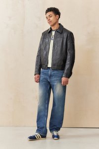 Black leather jacket with a collared neckline, white t-shirt, faded blue jeans, and blue sneakers with yellow stripes, standing against a wooden backdrop.