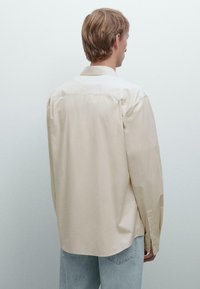 Beige lange mouwen shirt met een spitse kraag, knoopsluitingen aan de mouwen en een relaxte pasvorm. Glad materiaal met een lichte glans, met een achterste naaddetail.
