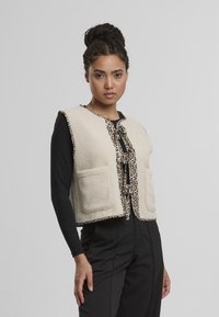 Beige fleecová vesta s leopardím potiskem, s páskem na zavázání a dvěma předními kapsami. Nositá přes černý dlouhý rukáv.