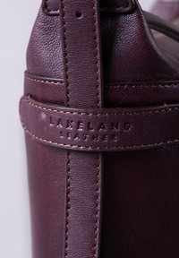 Burgundy leather bag with textured finish, featuring a secure strap and embossed "Lakeland Leather" logo, accentuated by contrasting stitching.