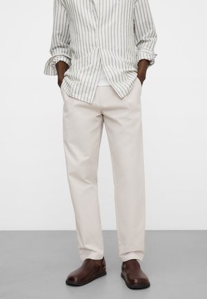 Uomo che indossa pantaloni beige dritti, scarpe marroni senza lacci e una camicia bianca con righe verticali verdi, mani nelle tasche.