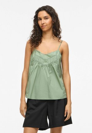 Nainen, jolla on kiharat hiukset, pukeutuneena vaaleanvihreään hihattomaan toppiin ja mustiin polvipitkiin shortseihin, seisoo yksinkertaisen taustan edessä.