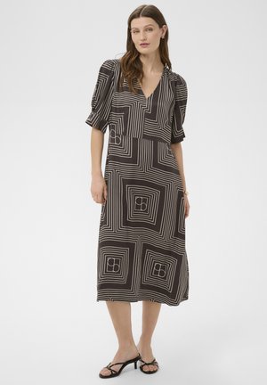 Kobieta ubrana w czarną sukienkę midi z białymi geometrycznymi wzorami kwadratów, z bufkami na krótkich rękawach, dekoltem w serek oraz czarnymi sandałkami na obcasie z paskami.
