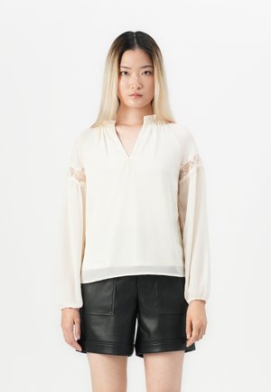 Giovane donna con lunghi capelli ombre, che indossa una blusa color crema con dettagli in pizzo e pantaloncini in pelle nera, in piedi contro uno sfondo bianco.
