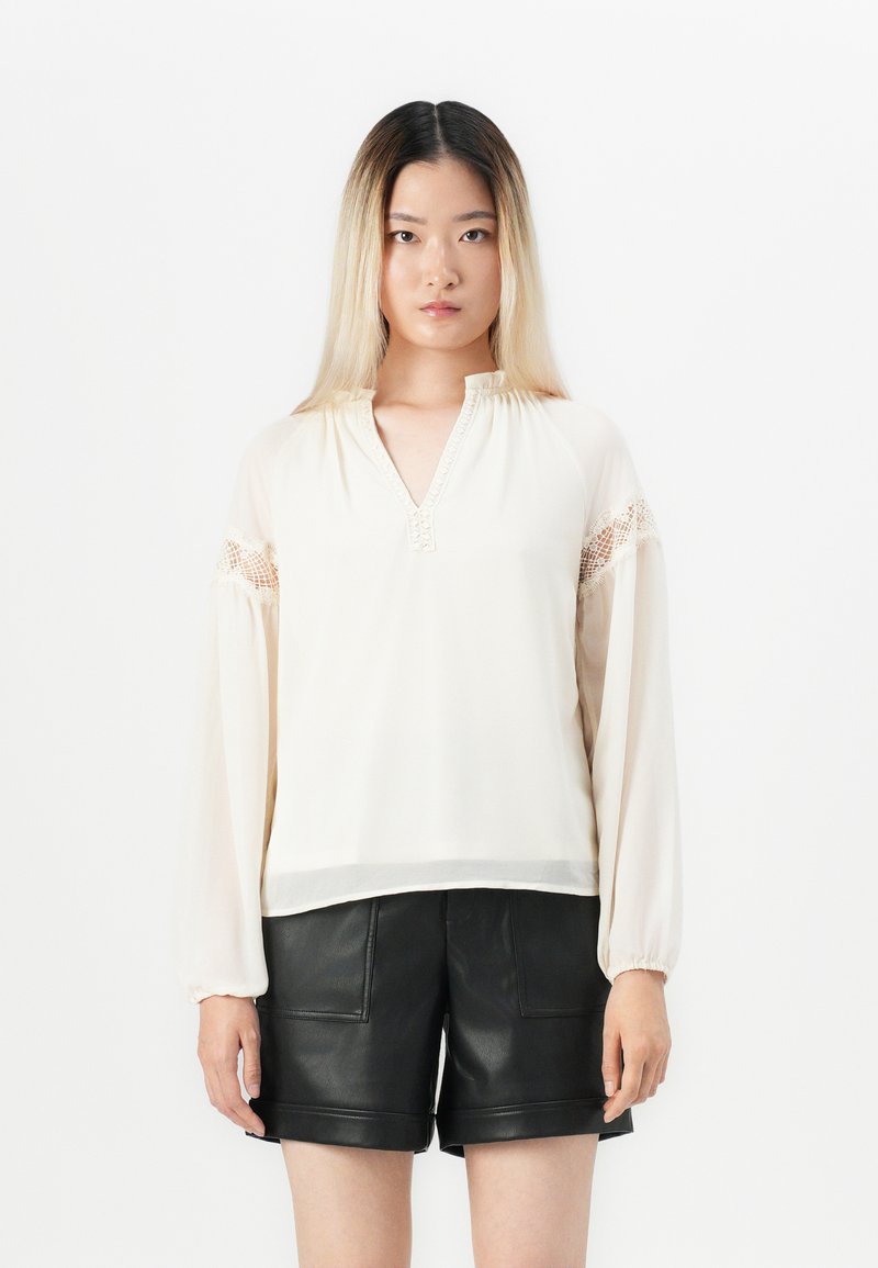 Giovane donna con lunghi capelli ombre, che indossa una blusa color crema con dettagli in pizzo e pantaloncini in pelle nera, in piedi contro uno sfondo bianco.