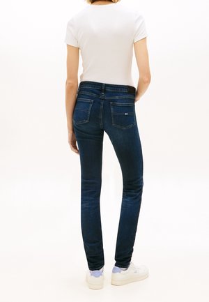 Žena v tmavě modrých úzkých džínách, bílém tričku s krátkým rukávem a bílých teniskách, stojí zády ke kameře.