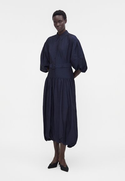 Γυναίκα που φοράει midi φόρεμα σκούρου ναυτικού μπλε με φουσκωτά μανίκια, ζώνη στη μέση και μαζεμένη φουσκωτή τελείωμα, συνδυασμένο με μαύρες μυτερές γόβες.