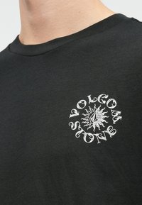 Zwarte katoenen t-shirt met een cirkelvormige witte grafiek met zon en geometrische ontwerpen. Klassieke ronde hals met korte mouwen.