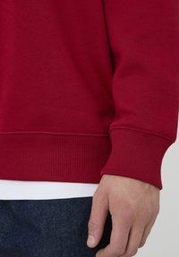 Sweat-shirt rouge avec poignets et ourlet côtelés, doté d'une texture douce. Une chemise blanche est portée en dessous, associée à un jean en denim foncé.