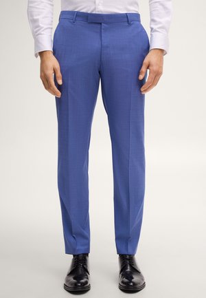 Mann trägt eng geschnittene blaue Anzughose, weißes Hemd und schwarze Anzugschuhe und steht vor einem schlichten hellen Hintergrund.