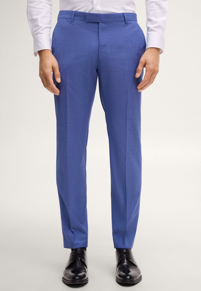 Man draagt nauwsluitende blauwe pantalon, wit overhemd en zwarte nette schoenen, staand tegen een effen lichte achtergrond.