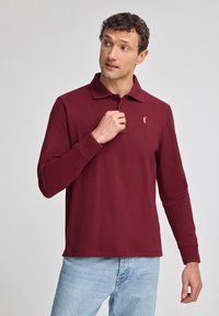 Bordeauxfarbenes Langarm-Polohemd mit Kragen, das ein dezentes Logo auf der linken Brust zeigt. Aus glattem Stoff gefertigt, kombiniert mit hellblauen Jeans.