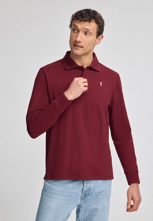 Bordeauxfarbenes Langarm-Polohemd mit Kragen, das ein dezentes Logo auf der linken Brust zeigt. Aus glattem Stoff gefertigt, kombiniert mit hellblauen Jeans.