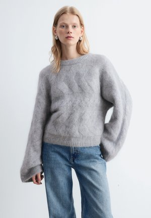 Femme aux cheveux blonds portant des créoles argentées, un pull gris ample en maille torsadée et un jean bleu, debout devant un fond clair.