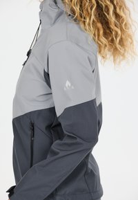 Personne aux cheveux bouclés portant une veste bicolore grise avec une poche latérale zippée et un logo réfléchissant sur la manche supérieure.