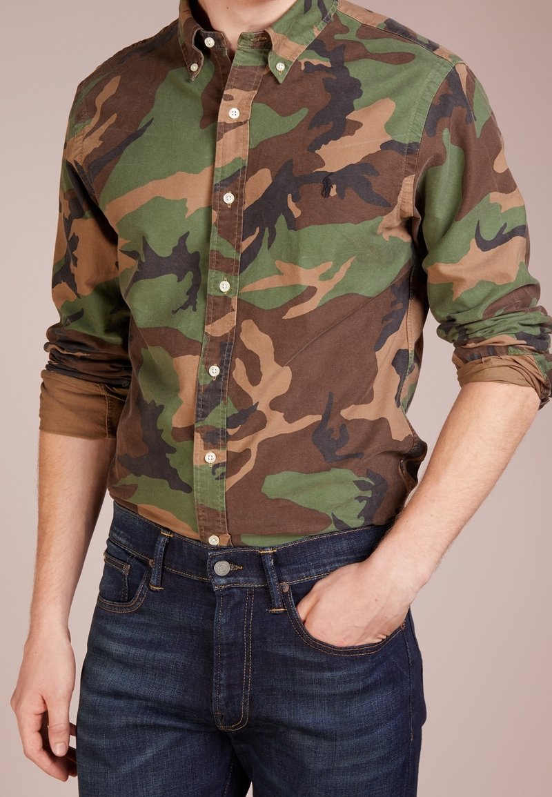 Camisa de botones de camuflaje verde y marrón en tela de algodón, con cuello y mangas enrolladas, combinada con jeans azul oscuro.