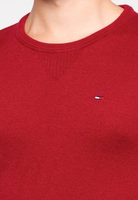 Pull en tricot rouge avec un col en côtes et un petit logo sur la poitrine. Le matériau semble doux et léger, avec une texture lisse.