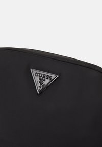 Sort nylon rygsæk med et triangel-farvet glossy Guess USA logo på fronten, med en glat tekstur og strømlinet design.