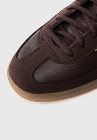 Κοντινό πλάνο σε μύτη αθλητικού παπουτσιού από σκούρο καφέ δέρμα και σουέτ με κορδόνια ίδιου χρώματος και διάφανο λαστιχένιο πάτο σε λευκό φόντο.