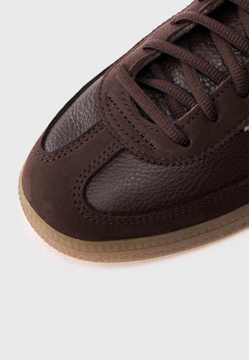 Nahaufnahme eines Sneaker-Vorderteils aus dunkelbraunem Leder und Wildleder mit passenden Schnürsenkeln und durchsichtiger Gummisohle vor weißem Hintergrund.