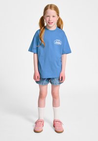 Blauwe t-shirt met korte mouwen met een logo op de borst, gecombineerd met denimshorts, witte kniekousen en roze sneakers.