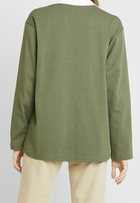 Personne portant un haut à manches longues vert olive avec un col blanc, associé à un pantalon beige, vue de dos.