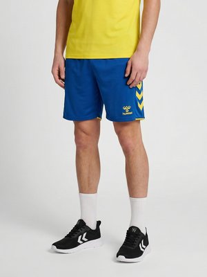 Mand iført blå sportsshorts med gule accenter, gul skjorte, hvide mellemlange sokker og sorte sportssko, stående på en ensfarvet baggrund.