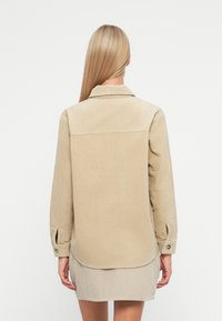 Μπεζ πουκάμισο από κοτλέ με διπλωμένο γιακά, μακριά μανίκια, κουμπωτές μανσέτες και στρογγυλή ραφή. Η πίσω όψη δείχνει έναν υφασμάτινο, ραβδωτό σχεδιασμό.