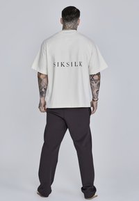 Krémové tričko s krátkym rukávom s potlačou "SIKSILK" v čiernej farbe na chrbte, kombinované s tmavými teplákmi. Vyrobené z mäkkého materiálu; voľný strih.