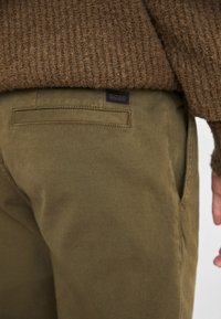 Olivgröna byxor i bomull med en texturerad yta. Har sidofickor och en marinblå logopatch på bakfickan.