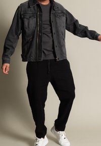 Man med mörk denimjacka, svarta joggingbyxor, grå t-shirt och vita sneakers, står med armarna lätt utsträckta.