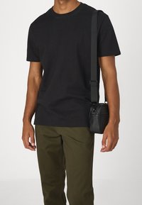 Svart t-shirt i bomull med rund hals, kombinerad med olivgröna byxor. En liten svart lädercrossbody-väska hänger från ett mörkt bälte.