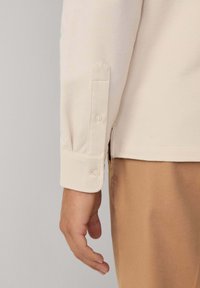 Ljus beige långärmad skjorta med knäppta manschetter, med mjuk textur och sidan slits detaljer, jämte bruna byxor.