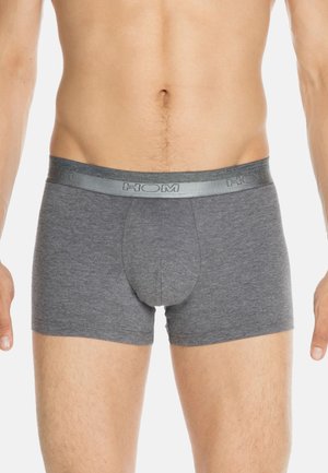 Homme portant un slip ajusté gris avec une ceinture de marque, se tenant contre un fond blanc, torse et parties supérieures des jambes visibles.
