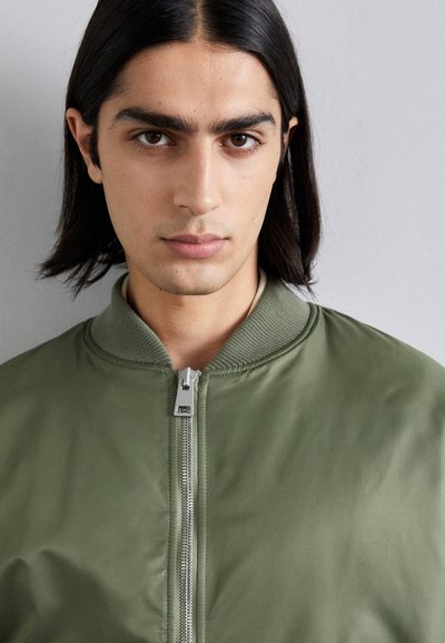 Chaqueta bomber en verde oliva, con cuello acanalado, cremallera frontal completa y textura de tela suave.