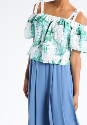 Vrouw draagt een witte off-shoulder top met groene bladmotieven en blauwe wijde, hoog getailleerde broek.