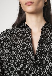 Schwarzes Shirt mit einem geometrischen weißen Quadratmuster, mit einem Kragen und einer Knopfdetaillierung an der Vorderseite. Glatte Textur.